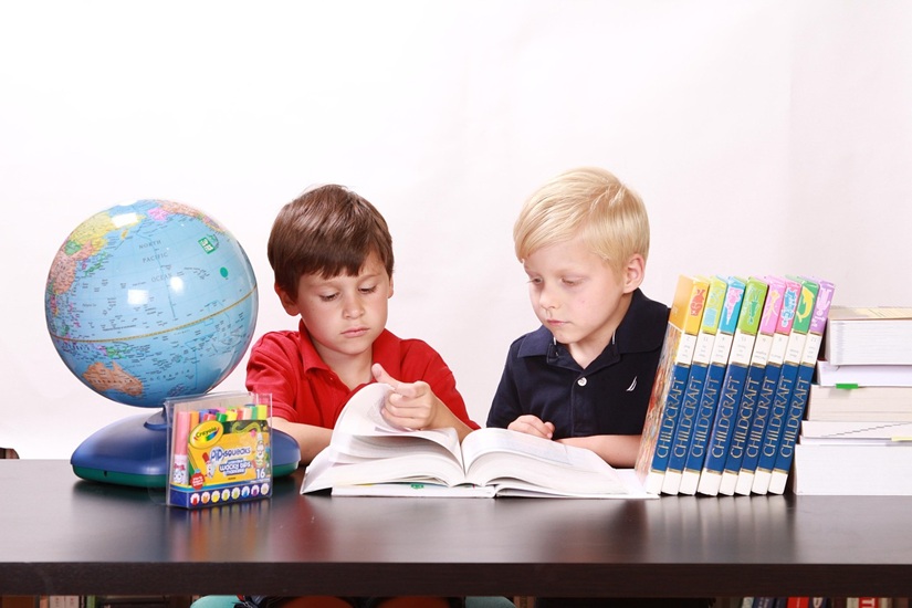 Two kids studying a book on a table with a globe and books