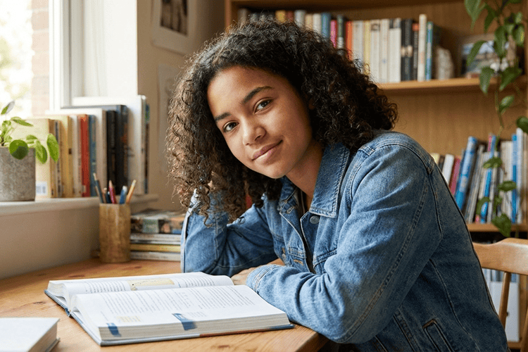 Confident teenage student sitting at desk with open book looking thoughtfully at camera in modern study space with natural lighting representing What are the 7 habits of a highly effective teenager